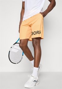 Orange athletic shorts with "BORG" printed in black, worn with a white t-shirt and white sneakers, holding a tennis racket.