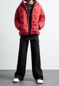 Czerwona kurtka puchowa z kapturem, zamkiem błyskawicznym i akcentem logo. W połączeniu z czarnymi spodniami o szerokich nogawkach i białymi trampkami.