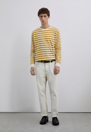 Joven de pie contra una pared lisa, vistiendo una camisa de manga larga a rayas amarillas y blancas, pantalones blanco sucio, cinturón negro y zapatos negros.