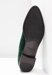 Semelle d'une chaussure habillée pour homme en daim vert foncé à bout pointu, reposant contre un mur blanc d'angle, montrant le talon noir et la semelle avec la taille 37.