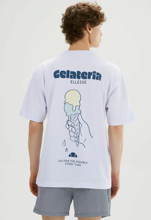 Joven de espaldas lleva una camiseta blanca con un gráfico de un cucurucho de helado doble derritiéndose y el texto "Gelateria Ellesse" y pantalones cortos a rayas.