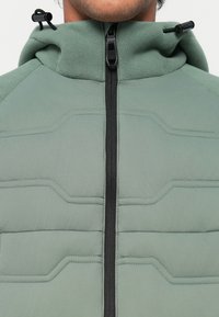 Veste en polaire verte avec un col haut, dotée d'une fermeture éclair à l'avant, d'un design texturé matelassé et de cordons réglables sur la capuche.