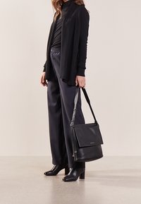 Borsa a tracolla in pelle nera con finitura texturizzata, chiusura a pattina e tracolla lunga. Indossata insieme a vestiti scuri e stivali con tacco a punta.