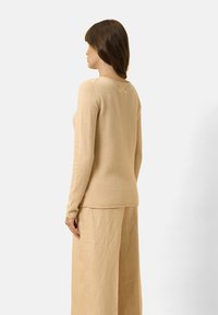 Beige Langarmpullover mit weichem Material, Rundhalsausschnitt und taillierter Passform. Kombiniert mit hellbeigen, weiten Hosen.