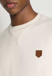 Světle béžová mikina s kulatým výstřihem, na hrudi má texturovanou hnědou koženou nášivku a viditelné švy.