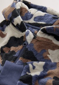 Sweat à capuche en polaire avec un motif camouflage en bleu, marron et crème. Il présente une texture douce avec des poignets côtelés et une fermeture éclair à l'avant.