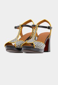 Μαύρα και κίτρινα πέδιλα με ψηλό τακούνι και γεωμετρικά σχέδια, ανοιχτή μύτη, λουράκι στον αστράγαλο και αντίθετη καφέ εσωτερική σόλα. Επίπεδη πλατφόρμα μπροστά.