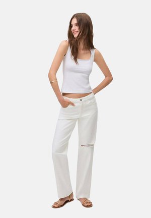 Vita denimbyxor med en avslappnad passform, som har ett slitet knä, tillsammans med en åtsittande vit linne. Modellen står med händerna i fickorna.