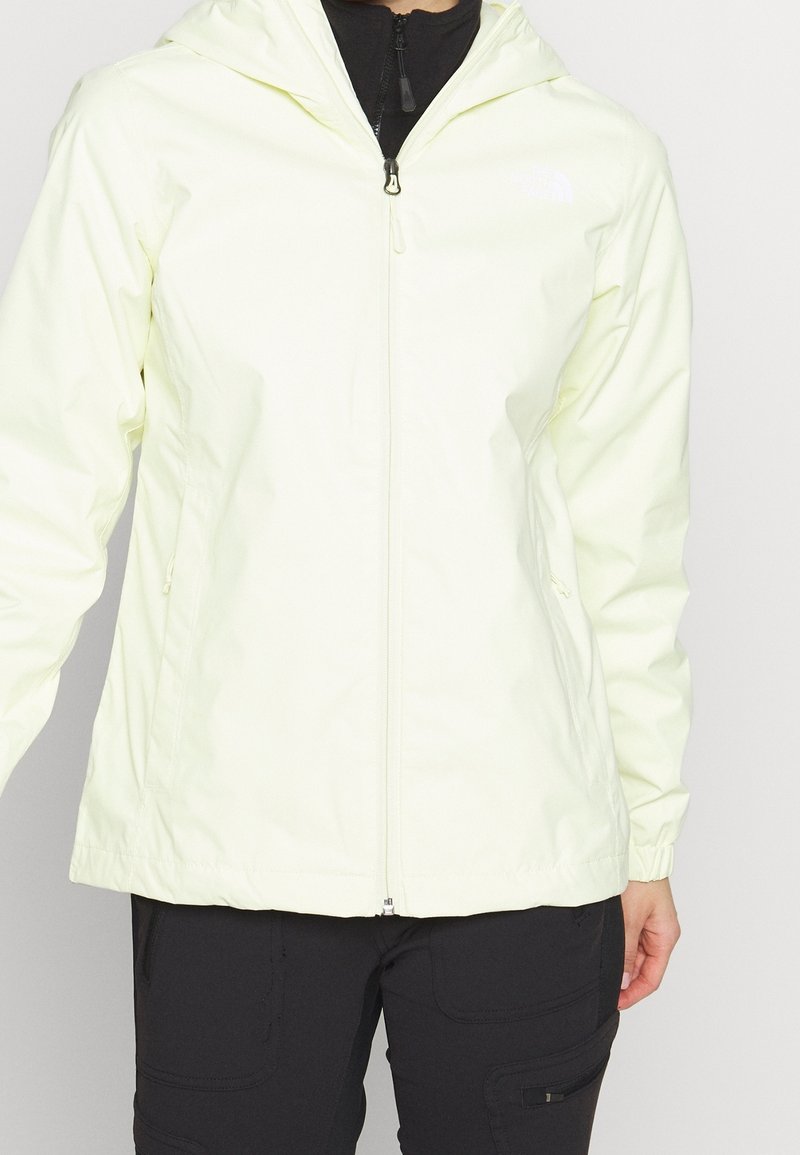 Personne portant une veste à capuche jaune pâle zippée sur un haut noir et un pantalon noir avec des poches zippées.