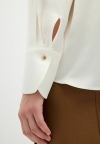 Camicia bianca con polsini abbottonati, caratterizzata da un dettaglio con fessura. Tessuto liscio con una leggera lucentezza, abbinata a un tessuto marrone sul bordo inferiore.