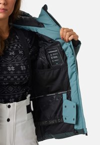 Eine türkisfarbene Außenjacke mit schwarzem Innenfutter zeigt ein Pflegeetikett. Das Design verfügt über Taschen und Belüftungsnetz und besteht aus wetterbeständigen Materialien.