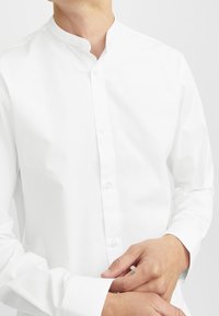 Vit skjorta med bandkrage, slät textur och långa ärmar. Har en subtil lyster med knappar längs frontplacket.