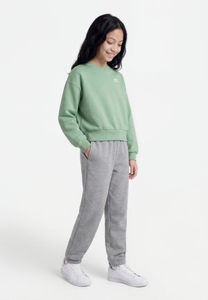 Ragazza sorridente con capelli lunghi e scuri indossa una felpa verde Nike, pantaloni della tuta grigi e scarpe da ginnastica bianche, in piedi con le mani in tasca.