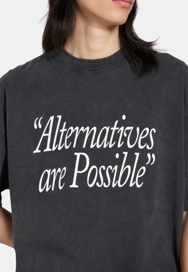 Personne portant un t-shirt gris foncé avec un texte blanc indiquant « Alternatives are Possible » sur le devant, sur un fond clair uni.