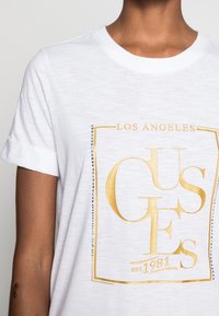 Bílý bavlněný tričko s ohrnutými rukávy, se zlatým grafickým nápisem "LOS ANGELES" a "EST. 1981", ozdobené stříbrnými nýty po straně.