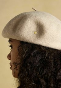 Gyapjú barett, világos bézs színű, lekerekített alakú, tetején hurokkal, kis arany díszítéssel. Puha textúrájú, sima felületű.