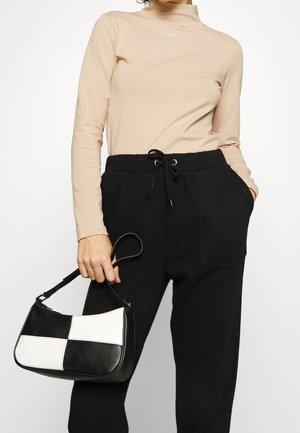 Frau trägt beige langärmeliges Oberteil und schwarze Hose mit Kordelzug, hält eine schwarz-weiß karierte Handtasche mit kurzem Henkel.