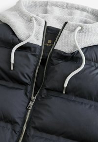 Doudoune noire avec une capuche grise, dotée d'une fermeture éclair argentée et de cordons de serrage, texture matelassée et étiquette logo à l'intérieur.