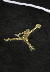 Sort stof med et guld broderet logo af en hoppefigur, der holder en bold, med en hvid accentlinje ovenfor. Glat tekstur.