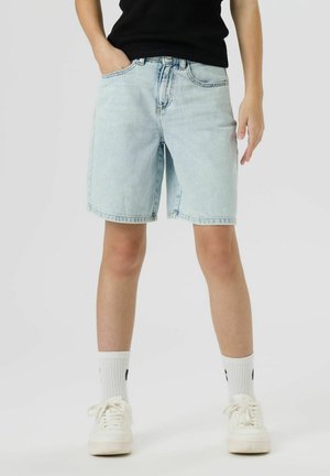 Noor inimene, kes kannab helesiniseid teksapükse, valgeid tenniseid ja valgeid sokke, seisab ühe käega taskus.