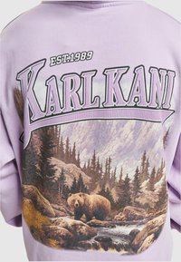 Felpa lavanda con un design grafico di un orso in un paesaggio forestale, con il testo "KARL KANI" in grassetto e colori a contrasto.