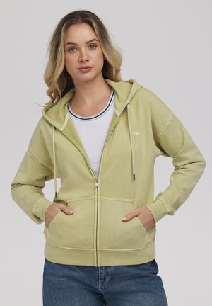 Kobieta ubrana w jasnozieloną bluzę z kapturem na zamek, na białej koszuli i niebieskich jeansach, stojąca z rękami w kieszeniach na jednolitym tle.