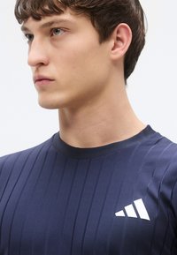 Eng anliegendes navyfarbenes Sportshirt mit geripptem Muster, versehen mit einem Rundhalsausschnitt und einem weißen Logo auf der linken Brust.