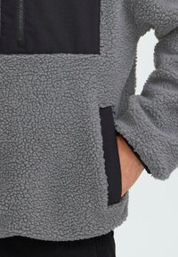 Grauer Fleece-Pullover mit schwarzem Stoffeinsatz und einer zipbaren Tasche. Die Textur ist weich und fluffig mit einer lässigen, kastigen Passform.