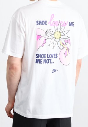 T-shirt en coton blanc avec un motif coloré au dos, comprenant du texte, des mains et un dessin de marguerite dans des tons rose, jaune et bleu.