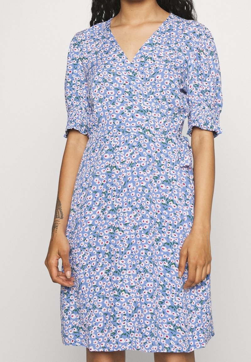 Femme portant une robe portefeuille bleu clair avec imprimé floral de marguerites blanches et roses et manches courtes bouffantes, debout devant un fond uni.
