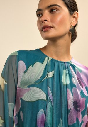 Transparent blågrön blus med blomstertryck i lila och gröna nyanser, med rynkad halsringning och mjukt, flytande tyg. Enkel, elegant design.