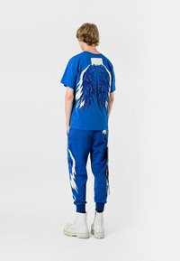 Camiseta de algodón azul con un diseño de alas en la parte posterior, combinada con joggers azules a juego que tienen acentos blancos. El modelo lleva botas blancas.