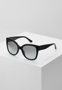 Óculos de sol oversized pretos com um acabamento brilhante e lentes gradientes, apresentando armações angulares e um design elegante. Acentuados com a marca "vogue".
