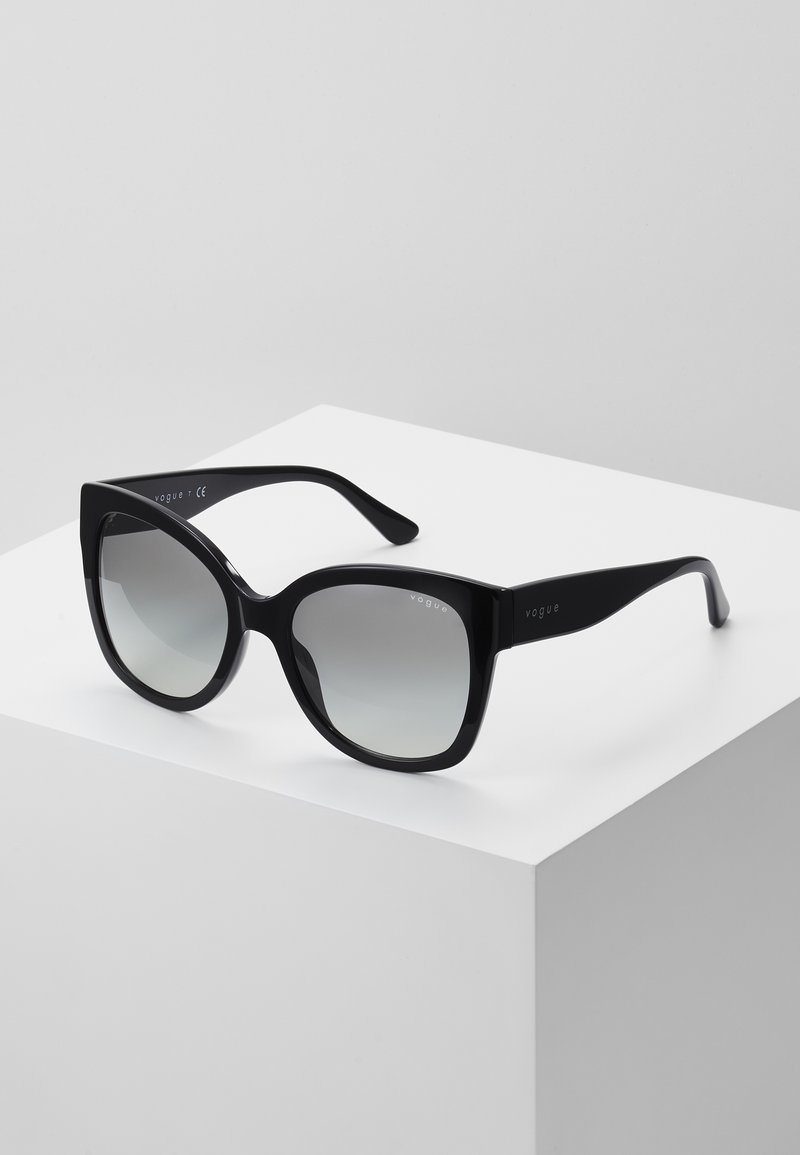 Óculos de sol oversized pretos com um acabamento brilhante e lentes gradientes, apresentando armações angulares e um design elegante. Acentuados com a marca "vogue".