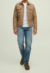 Brun bomullsjacka med två bröstfickor, vit t-shirt under, ljusblå jeans och bruna skor med snören. Avslappnad passform och utseende.
