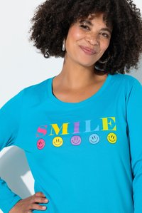 Türkisfarbenes Langarmshirt mit mehrfarbigem "SMILE"-Text und Smiley-Grafiken. Weiche Baumwollmischung, runder Ausschnitt, lockere Passform.