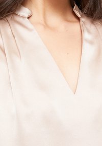 Ljusrosa blus med V-ringning och kort, mjuk krage. Tyget har en slät, satinliknande textur med subtila veck nära ringningen.