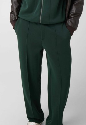 Personne portant un pantalon vert foncé et un haut à fermeture éclair assorti, mains dans les poches, manches d'une veste en cuir marron, et chaussures blanches visibles.