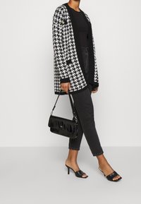 Houndstooth cardigan in zwart en wit, gebreide textuur, open voorkant, gecombineerd met zwarte fitted jeans, zwarte hakken mules en een zwarte handtas.