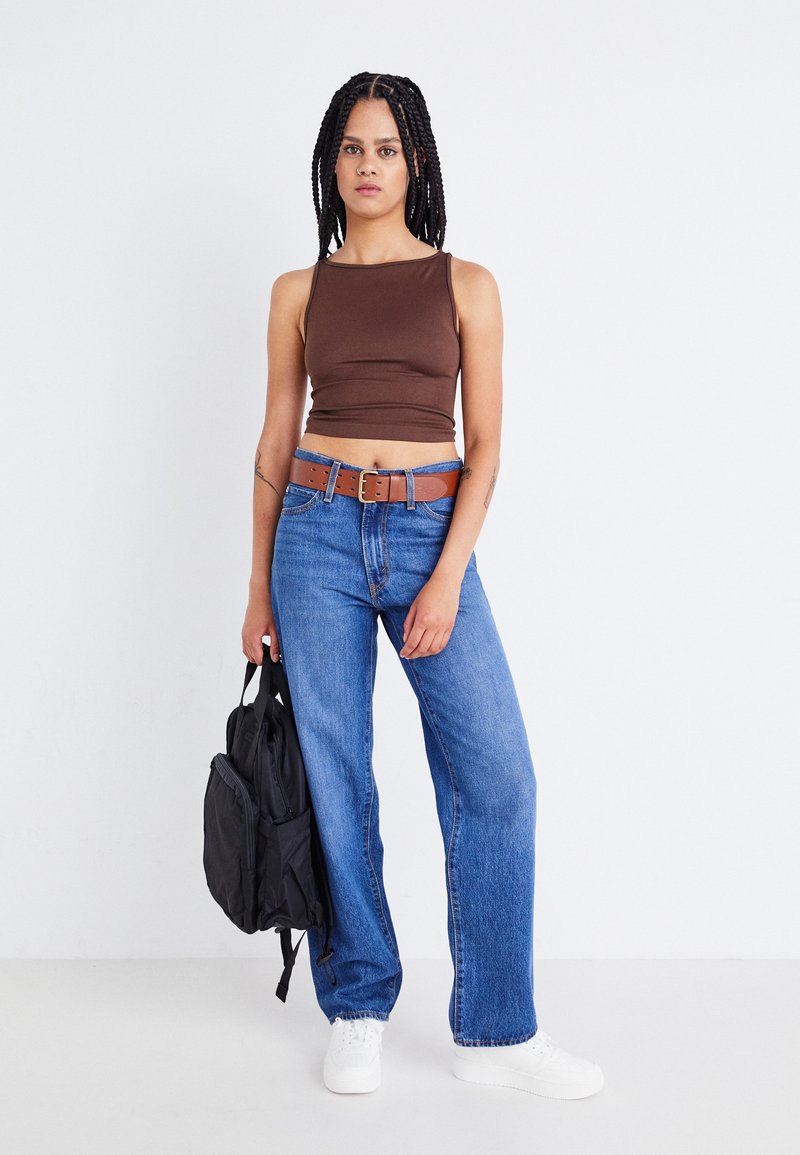 Brun tætsiddende crop-top, blå flagrende jeans med et brunt bælte og hvide sneakers. Sort ryksæk holdt i den ene hånd foran en ensfarvet baggrund.