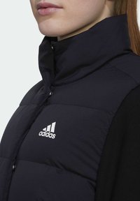 Svart vadderad väst med hög krage, med vitt Adidas-logotyp, tryckknappar och slät textur. Närbild av nacklinjen och bröstområdet.