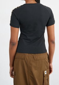 Person som bär en svart kortärmad skjorta med mönstrade axelränder och en högmidjad brun cargoskirt med klafffickor, stående mot en vit bakgrund.