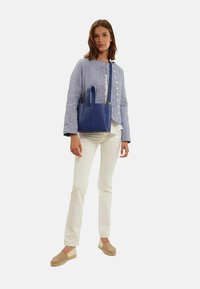 Sac à main en cuir bleu avec des poignées courtes, porté avec une veste en lin gris clair et un pantalon blanc, présentant une silhouette décontractée et ajustée.