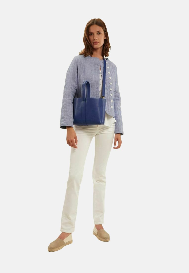 Sac à main en cuir bleu avec des poignées courtes, porté avec une veste en lin gris clair et un pantalon blanc, présentant une silhouette décontractée et ajustée.