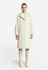 Lång, krämfärgad ullkappa med stor krage och ballongärmar, med sidofickor och rak skärning. Kombinerad med knähöga beige stövlar.