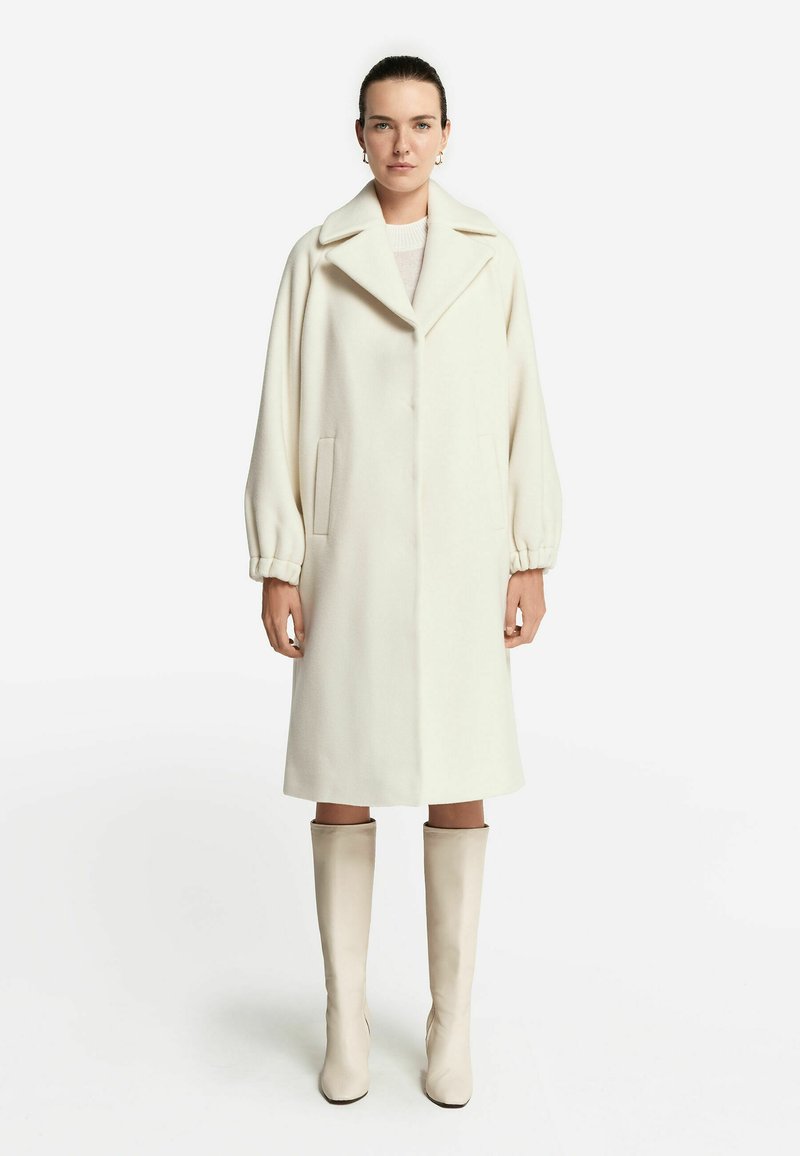 Lång, krämfärgad ullkappa med stor krage och ballongärmar, med sidofickor och rak skärning. Kombinerad med knähöga beige stövlar.