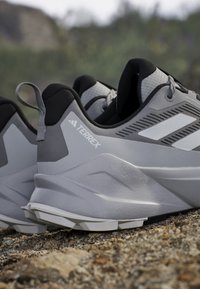 Zapatillas deportivas grises con un upper texturizado, talón reforzado y detalles en blanco. El diseño incluye una tira de agarre y una suela curva.