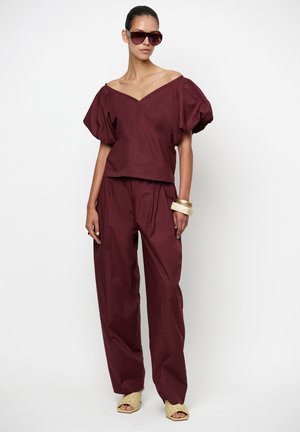 Vrouw draagt een donker bordeauxrode off-shoulder top met pofmouwen en bijpassende wijde broek, met beige geweven sandalen en grote bruine zonnebrillen.