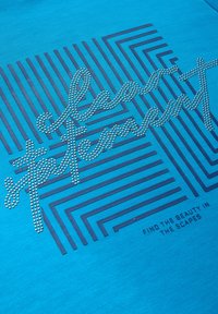 Türkises T-Shirt mit einem Design aus silbernen Nieten, die in einer geschwungenen Schrift angeordnet sind, kombiniert mit marineblauen geometrischen Linien und Text darunter.
