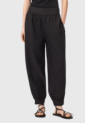 Vrouw draagt losse zwarte broek met ingesnoerde boorden en zwarte sandalen met bandjes en platte zolen, staand tegen een effen lichte achtergrond.